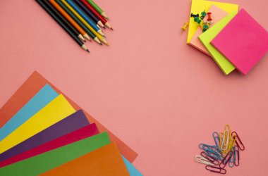 Kalemler, çok renkli kağıtlar ve pembe arkaplanda ataçlar.