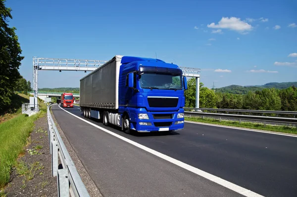 Blå lastbil passerar genom elektroniska toll gates på motorvägen i en trädbevuxen landskap. Röd lastbil, bridge och skogklädda berg i bakgrunden. Vita moln i den blå himlen. Stockbild