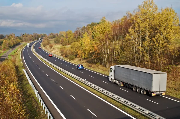 Asfalt dálnice s bílými kamiony a červené osobních automobilů v malebnou podzimní krajinou. Slunečný den s ponurý mraky na pozadí. Pohled shora. — Stock fotografie