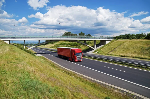 Röd lastbil på motorvägen går under betongbro på landsbygden. Visa från ovan. Solig dag med blå himmel och vita moln. — Stockfoto