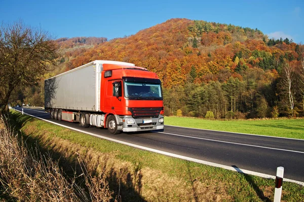 Camion rosso su strada asfaltata sotto montagna boscosa di colori autunnali incandescenti. Chiaro giorno di sole con cieli blu . — Foto Stock