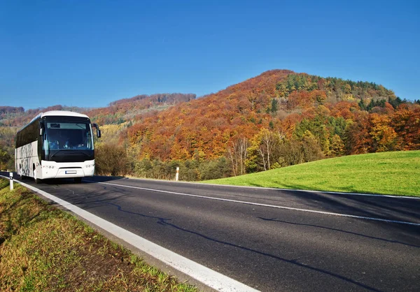 Vit buss anländer till asfaltvägen genom dalen nedanför det skogsbevuxna bergen av glödande höstfärger. Klar solig dag med blå himmel. Stockfoto