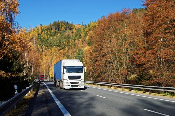 LKWs auf Asphaltstraße in einem bewaldeten Tal unterhalb des Berges, die in herbstlichen Farben lodern. sonniger Herbsttag mit blauem Himmel. — Stockfoto