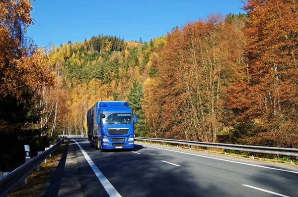 Camion bleu sur route asphaltée dans une vallée boisée sous la montagne, flamboyant de couleurs automnales. Journée ensoleillée d'automne avec ciel bleu . Photos De Stock Libres De Droits