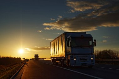 Gün batımında kırsal manzara içinde asfalt karayolu üzerinde kamyon. Günbatımı renkleri mavi gökyüzünde bulutlar.
