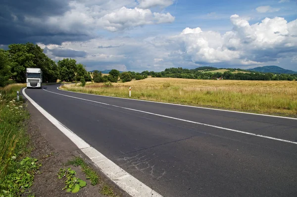 El camino de asfalto en el campo rural con nubes dramáticas en el cielo. Camión blanco salir de la curva entre los árboles en la distancia. Campo de maíz y montañas boscosas en el fondo . — Foto de Stock