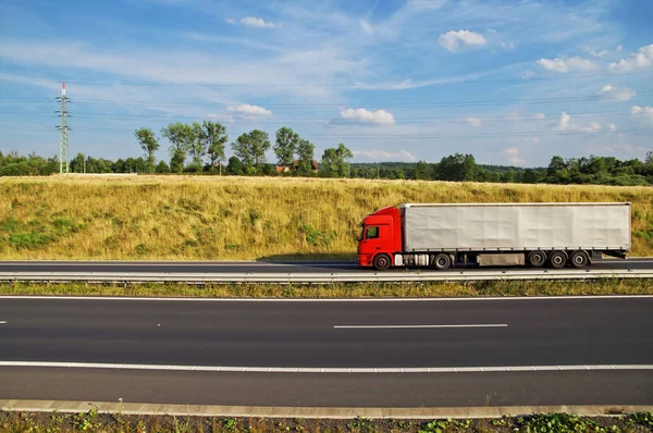 Roter LKW, der auf der asphaltierten Autobahn unter dem grasbewachsenen Hang fährt. Strommasten, Wald- und Dorfhäuser am Horizont. sonniger Sommertag mit blauem Himmel und weißen Wolken. — Stockfoto