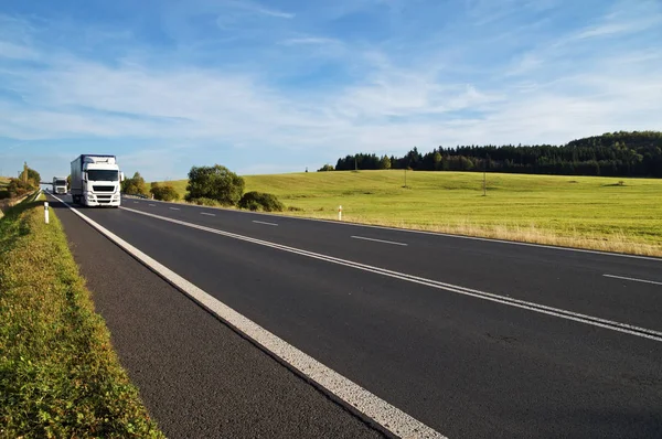 Asphalt road rising to the horizon in rural landscape. White trucks arriving from afar. Meadow and forest in the background. Sunny day with blue skies and white clouds. Royalty Free Stock Images