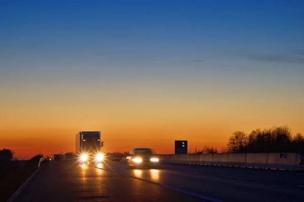 Highway with oncoming trucks and a car after sunset. Shining the spotlight cars. Information boards about the temperature. Blue and orange sky is clear. — Stock Photo, Image