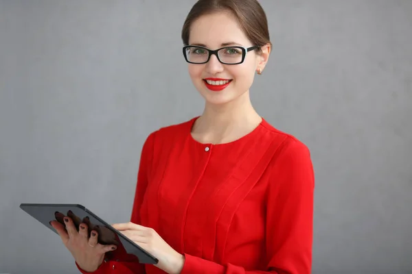 Zakenvrouw in een rood shirt en glazen houden een tablet in zijn handen op een grijze achtergrond — Stockfoto