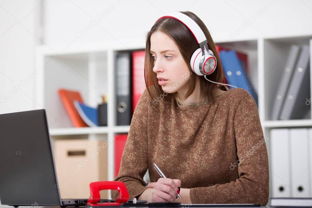 Beautiful female student with headphones listening to music and learning. Hold the handle in his hand and looking at laptop monitor