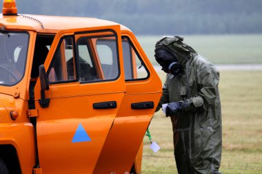 Gaz maskeli adam ve turuncu arabanın yanındaki güvenlik elbisesi.
