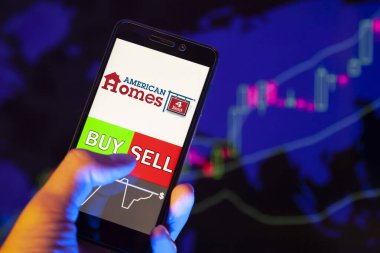 YESSENTUKI, RUSSIA - July 25, 2019: Company logo American Homes 4 Rent on smartphone screen, hand of trader holding mobile phone showing BUY or SELL on background of stock chart
