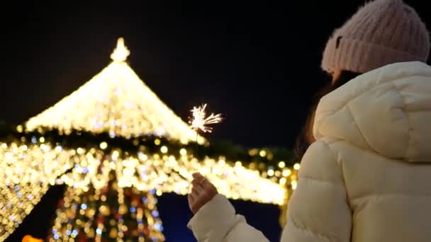 Giovane donna in cappotto e cappello con un Sparkler vicino all'albero di Natale. Vista da dietro — Video Stock