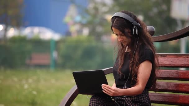 Giovane donna su una panchina con cuffie e laptop che ascolta musica all'aperto — Video Stock