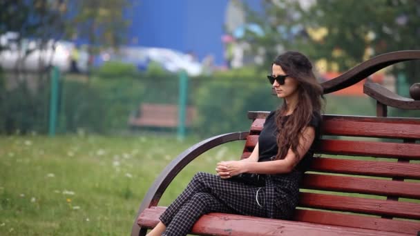 Jeune femme en lunettes de soleil avec les cheveux longs assis sur un banc dans un parc et triste — Video