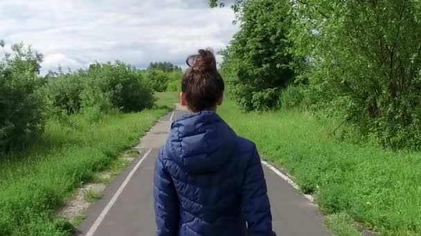 Mujer joven en chaqueta azul camina a lo largo del camino en verano Park — Vídeo de stock