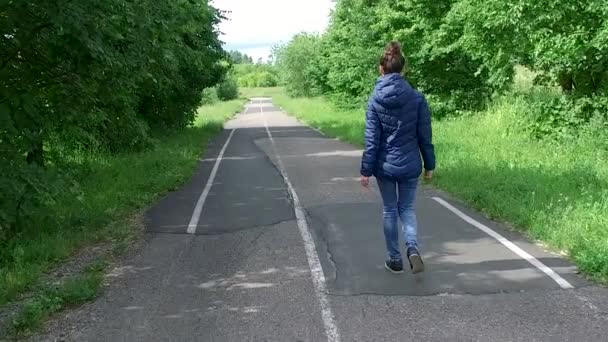 Young woman in blue jacket walks along path in summer Park on cool day — Stock Video