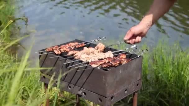 Grill with barbecue on background of body of water, human hand rotates skewers — Stock Video