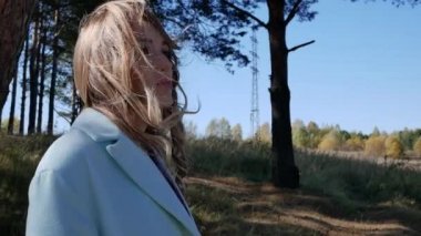 Mavi paltolu güzel bir kadın ve rüzgarlı bir havada sonbahar ormanında saçlarını örüyor.