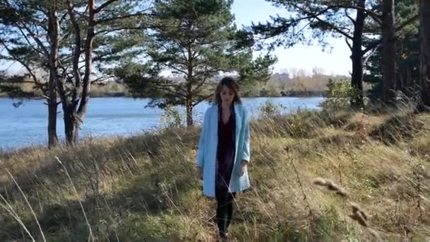 Piękna młoda kobieta w niebieskim płaszczu spacerująca ścieżką w jesiennym lesie przy wietrznej pogodzie — Wideo stockowe
