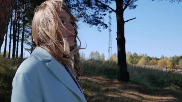 Hermosa mujer joven con abrigo azul y se mete el pelo en el bosque de otoño en tiempo ventoso — Vídeos de Stock