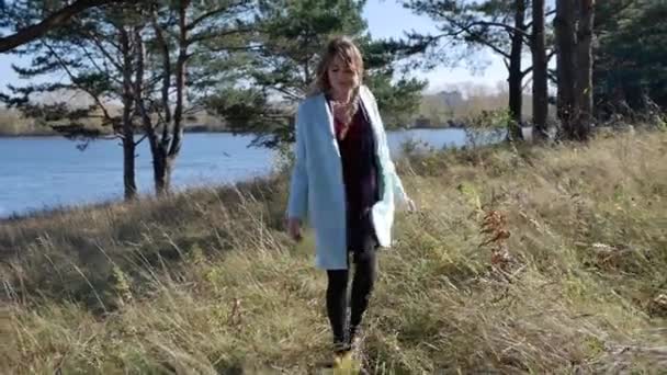 Piękna młoda kobieta w niebieskim płaszczu spacerująca ścieżką w jesiennym lesie przy wietrznej pogodzie — Wideo stockowe