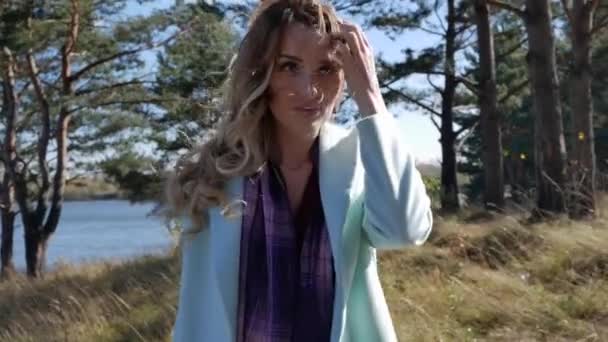 Hermosa joven con abrigo azul caminando por el camino en el bosque de otoño en tiempo ventoso — Vídeos de Stock