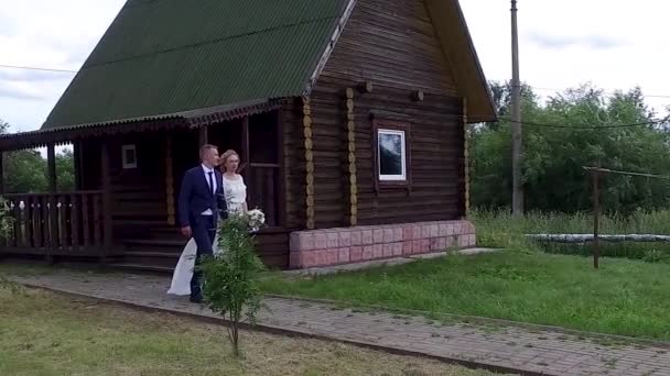 Beautiful newlyweds bride and groom with bouquet of flowers go happy holding hands on background of country house — Stok video