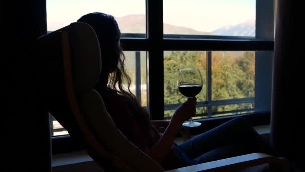 Силуэт женщины в кресле-качалке с бокалом вина в руке на фоне окна с красивым пейзажем гор — стоковое видео