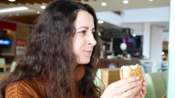 Joven hermosa mujer sonriente comiendo una hamburguesa en la cafetería — Vídeo de stock