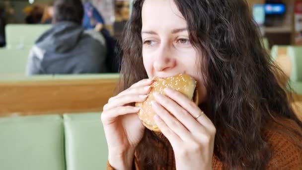 Jovem bela mulher sorridente comendo um hambúrguer no café — Vídeo de Stock