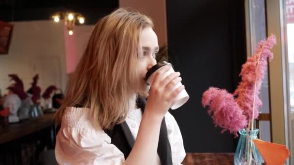 Blonde jeune femme dans café boire du café à partir d'une tasse de papier et regarder par la fenêtre — Video