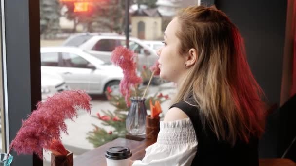 Молода жінка в кафе дивиться у вікно, чекає когось і п'є каву — стокове відео