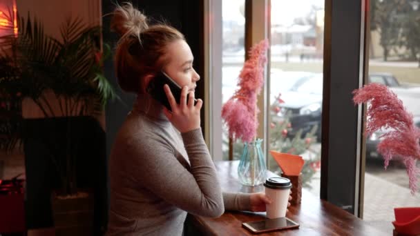 カフェで電話で話している若いブロンドの女性は、紙コップから笑顔で笑顔とコーヒーを飲んでいます。彼女は窓の外を見ている人を待っている — ストック動画