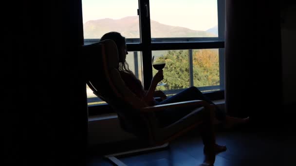 Silueta mladé krásné ženy v houpacím křesle se sklenkou vína na pozadí okna s krásnou krajinou hor — Stock video