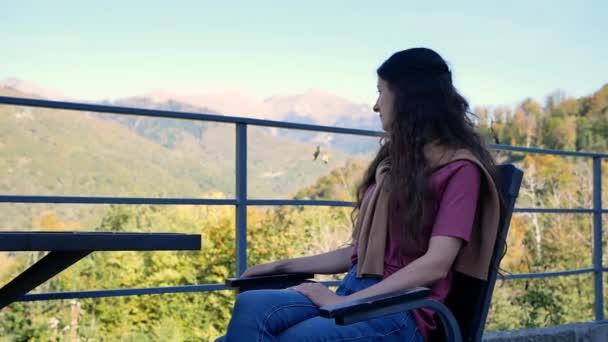 Hermosa joven mujer disfrutando de hermosos paisajes de montañas mientras se sienta a la mesa en la cafetería al aire libre — Vídeos de Stock