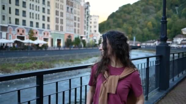 Beautiful young woman walking along mountain river in city at sunset — Stock Video