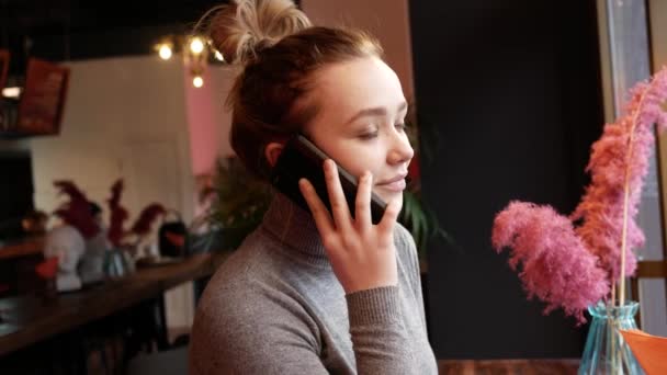 Fiatal szőke nő beszél telefonon egy kávézóban, nevet és mosolyog, és iszik kávét papír kupa — Stock videók