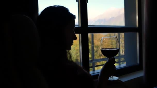 Силуэт молодой красивой женщины с бокалом вина в кресле-качалке на фоне окна с красивым пейзажем гор — стоковое видео