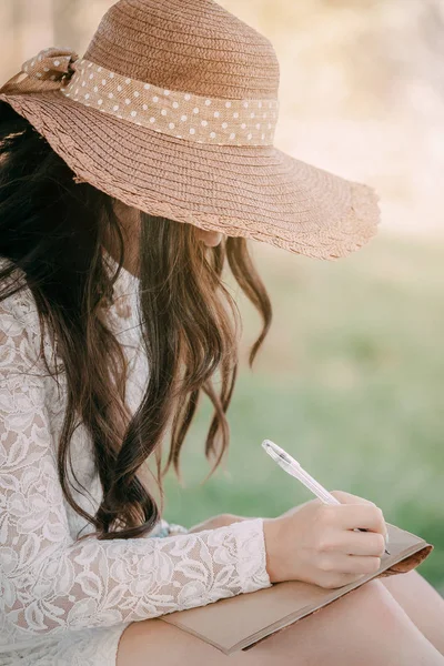 young woman in a straw hat with long hair sits on the shore of the lake on the grass and writes a letter. concept of romance