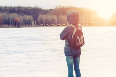 a man with a backpack, young woman in a blue jacket and hood looks into the distance at a frozen lake