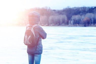 a man with a backpack, young woman in a blue jacket and hood looks into the distance at a frozen lake