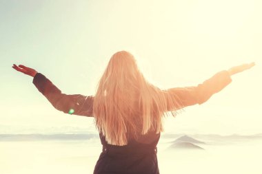 A young blond woman with raised hands enjoys the fresh mountain air and beautiful views of the mountains.