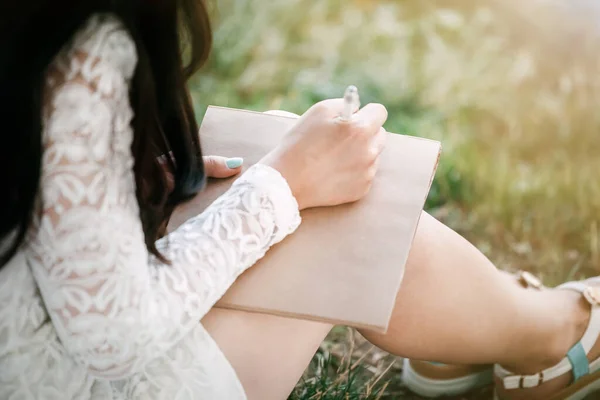 young woman in a straw hat with long hair sits on the shore of the lake on the grass and writes a letter. concept of romance