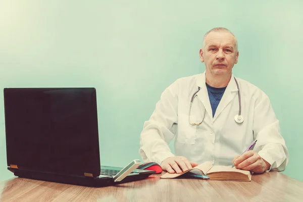 medicine profession technology and people concept smiling male doctor sitting at table with laptop and writes in the diary