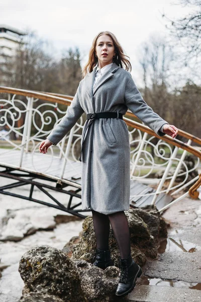 아름다운 다리 근처의 회색 코트를 입고 공원에서 포즈를 취하는 여자 모델 — 스톡 사진