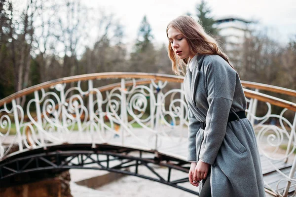 Girl model posing in park in a gray coat near a beautiful bridge — Stock Photo, Image