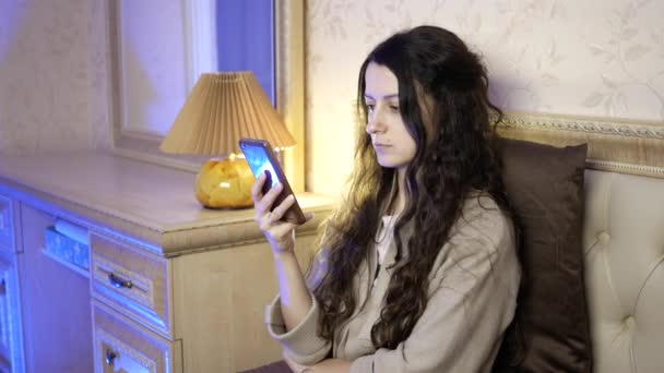Красива молода жінка використовує мобільний телефон, сидячи на ліжку в спальні — стокове відео