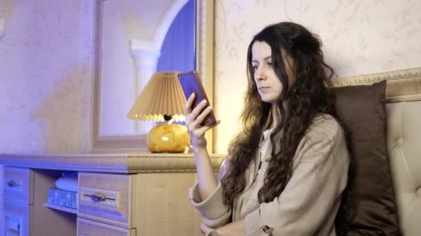 Schöne junge Frau benutzt Handy, während sie im Schlafzimmer auf dem Bett sitzt — Stockvideo